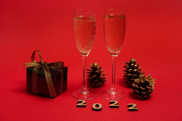 Een compositie van nieuwjaarsobject, houten 2022 cijfers, gouden dennenappels, kerstcadeau in glitter inpakpapier met gouden strik, champagnefluiten met mousserende wijn op rode achtergrond, kopieer ruimte