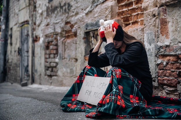 Een dakloze vrouw zit op straat in de schaduw van een huis en vraagt om hulp en geld Problemen van grote moderne steden Pak je hoofd vast met je handen Armoededrugs
