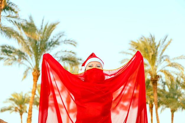 Een danseres van oosterse dans in een kerstmuts op een achtergrond van palmbomen met een gesloten cape over haar gezicht.