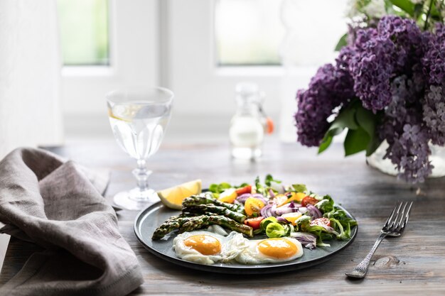 Een deel van veldsla met rode uien, gele tomaten, asperges en twee eieren op een plaat op een houten tafel.