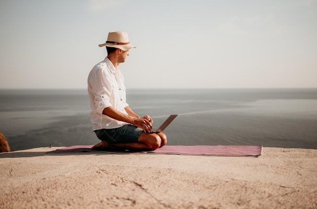 Een digitale nomade een man met een hoed een zakenman met een laptop doet yoga op de rotsen bij de zee bij