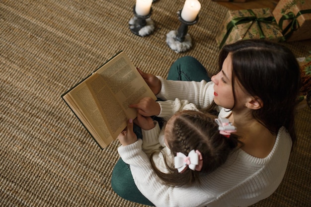 Een dochtertje en moeder in witte truien lezen een boek bij de kerstboom. bovenaanzicht