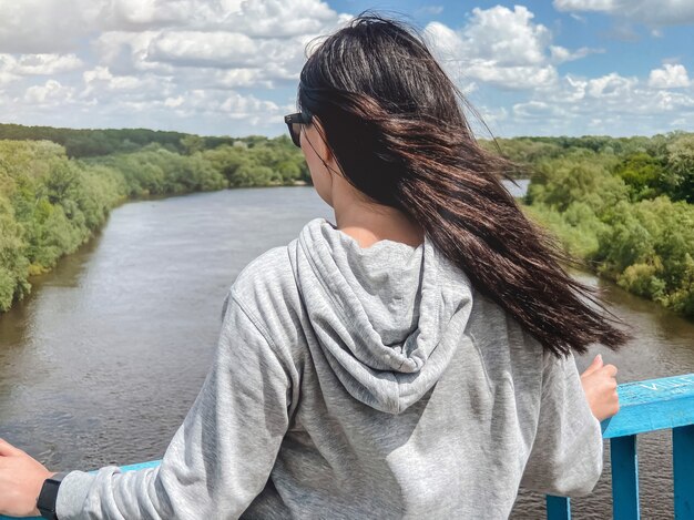 Een donkerharig aukasisch meisje staat met haar rug in een lichtgrijs jasje en kijkt naar de rivier