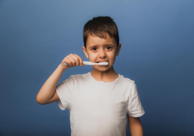 Een donkerharige jongen in een wit T-shirt op een blauwe achtergrond poetst zijn tanden met een tandenborstel. baby hygiëne