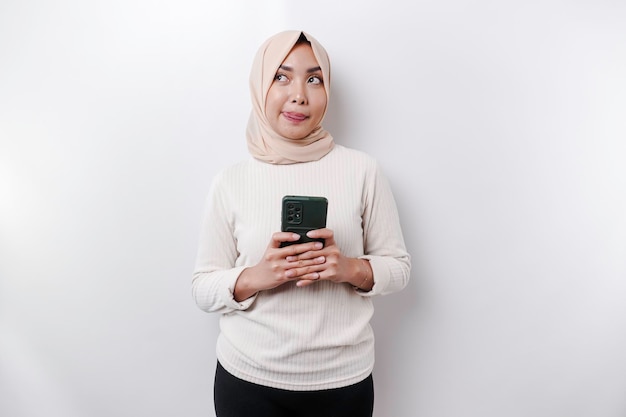 Een doordachte jonge Aziatische moslimvrouw die een hijab draagt en haar kin vasthoudt terwijl ze telefoneert geïsoleerd door een witte achtergrond