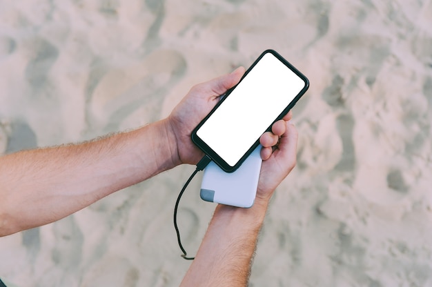 Een draagbare oplader in de handen van een man op de ruimte van strandzand. powerbank laadt de telefoon op.