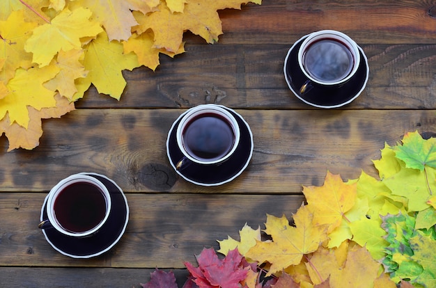 Een drie kopjes thee onder een set van vergeling gevallen herfstbladeren op een achtergrond oppervlak van natuurlijke houten planken van donker bruine kleur