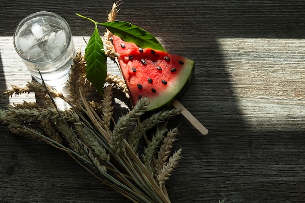 Een driehoekig stuk zoete watermeloen op een ijsje, een koud drankje met ijs en rijpe tarweoren op een houten oppervlak