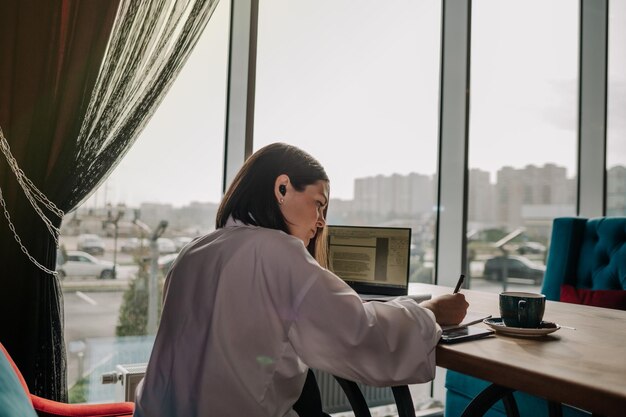 Een drukke brunette vrouw in een wit overhemd zit aan een tafel in een café en werkt op een laptop