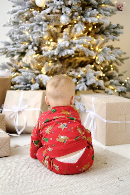 Een eenjarig kind in pyjama met een kerstprint zit op de grond en kijkt naar een stapel geschenken onder de kerstboom