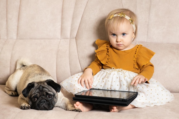 Een eenjarig meisje met een mopshond kijkt tekenfilms op een tablet terwijl ze op de bank zit Gadgets voor kinderen schermtijd