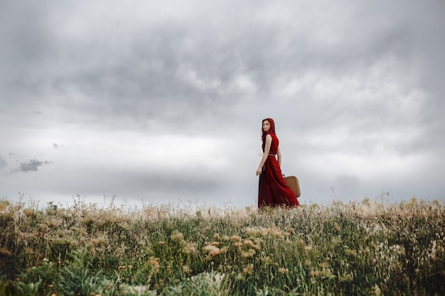 Een eenzaam rood haarmeisje in rode kleding met een vintage koffer en wandelen in de natuur