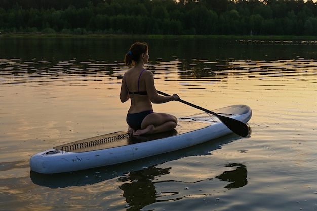 Een eenzame vrouw rijdt op een sup board op een meer omringd door gras en dicht groen bos bij zonsondergang. Lake Lebyazhye, Kazan.