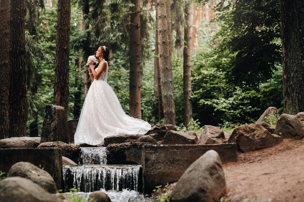 Een elegante bruid in een witte jurk en handschoenen met een boeket staat bij een beekje in het bos, genietend van de natuur. een model in een trouwjurk en handschoenen in een natuurpark. wit-rusland