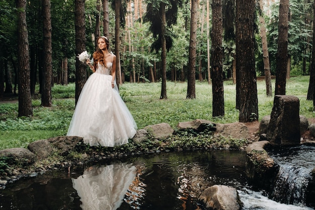 Een elegante bruid in een witte jurk en handschoenen met een boeket staat bij een beekje in het bos, genietend van de natuur. Een model in een trouwjurk en handschoenen in een natuurpark. Wit-Rusland
