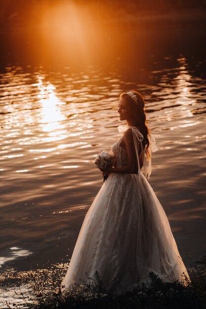 Een elegante bruid in een witte jurk en handschoenen staat bij de rivier in het park met een boeket, genietend van de natuur bij zonsondergang. Een model in een trouwjurk en handschoenen in een natuurpark. Wit-Rusland