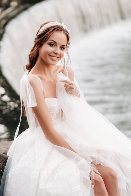 Een elegante bruid in een witte jurk, handschoenen en blote voeten zit in de buurt van een waterval in het park en geniet van de natuur. Een model in een trouwjurk en handschoenen in een natuurpark. Wit-Rusland