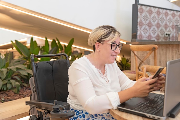 Een elegante vrouw met een handicap die in een rolstoel in een coffeeshop zit en berichten op haar mobiele telefoon checkt