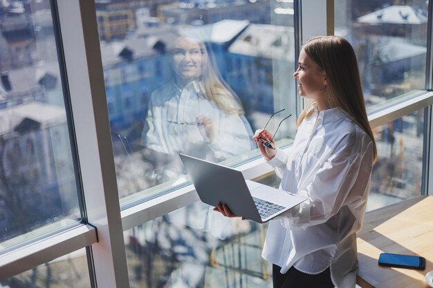 Een elegante vrouwelijke zakenvrouw met een bril voor oogcorrectie houdt een moderne laptop in haar handen bij een raam in een modern kantoor