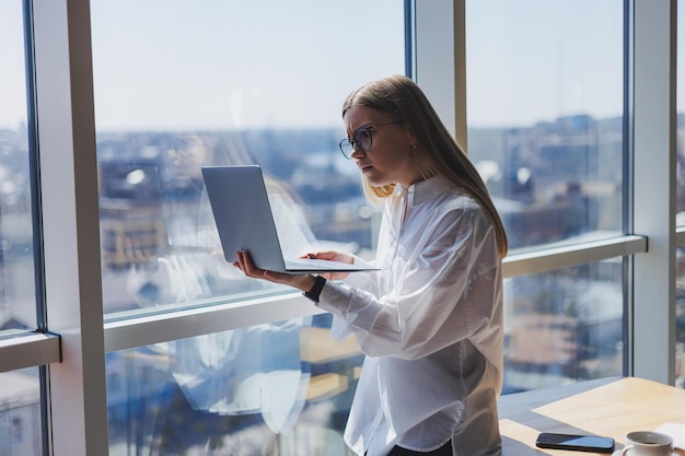 Een elegante vrouwelijke zakenvrouw met een bril voor oogcorrectie houdt een moderne laptop in haar handen bij een raam in een modern kantoor
