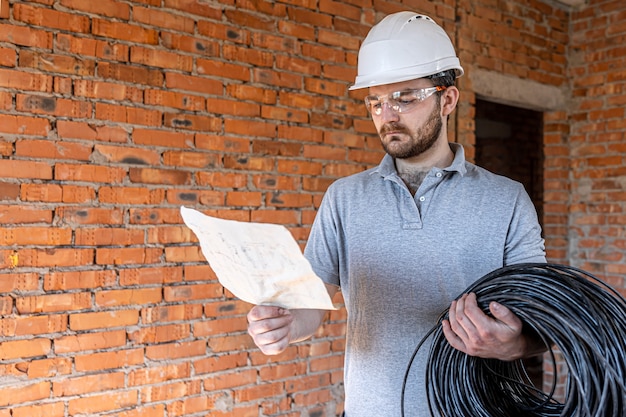 Een elektricien onderzoekt een constructietekening terwijl hij een elektrische kabel in zijn hand houdt op een werkplek.