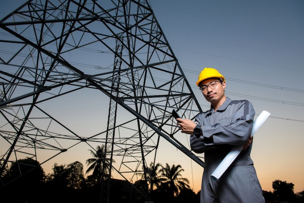 Een elektrotechnisch ingenieur die gele hoed draagt ​​houdt zwarte mobiele telefoon bij elektriciteitsmacht pol