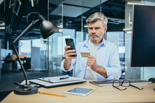 Een ervaren peinzende zakenman kiest ervoor om in een online winkel te winkelen, gebruikt de applicatie op de telefoon, een man werkt op kantoor op het werk