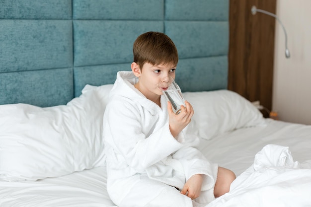 Een Europees kind houdt een glas schoon water vast Een jongen in een witte jas zit op het bed met een glas water