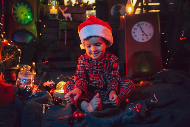 Een fantastische kerstsfeer. Gelukkig jongetje in mooie pyjama's en kerstmuts in kerstversiering. gekleurd licht. foto voor een ansichtkaart.