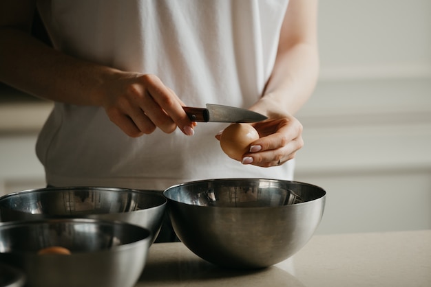 Een foto van de handen van een jonge vrouw die de eierschaal breekt met het mes boven de roestvrijstalen kom in de keuken