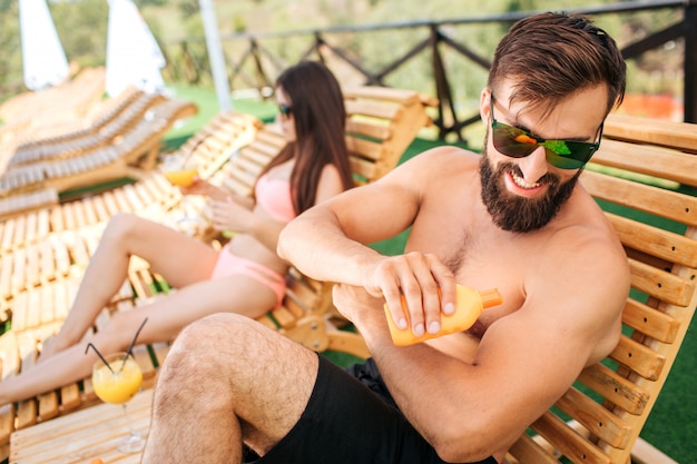 Een foto van een bebaarde man zittend op een zonnebank en wat zonwerende crème op de huid van een oranje fles. Hij draagt een zonnebril. Guy lacht. Meisje zit naast hem. Ze houdt een glas cocktail vast.