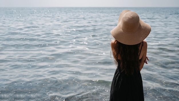 Een foto van een mooie jonge vrouw met lang donker haar in een jurk en een strohoed die op het strand poseert en met haar rug naar de eindeloze zee kijkt Een plek voor je tekst