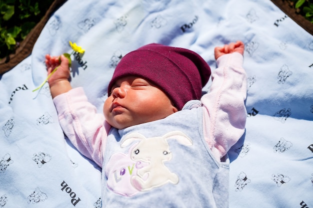 Een fotoportret van een baby met een rode dop ligt op een witte plaid op groen gras. De lente is op straat, de zon schijnt op het kind