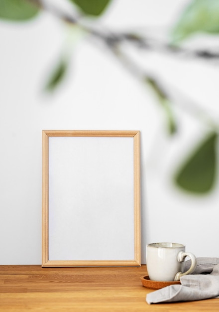 Een frame met een leeg doek tegen een lichte muur en op een houten tafel met vaag groen gebladerte en een kop thee