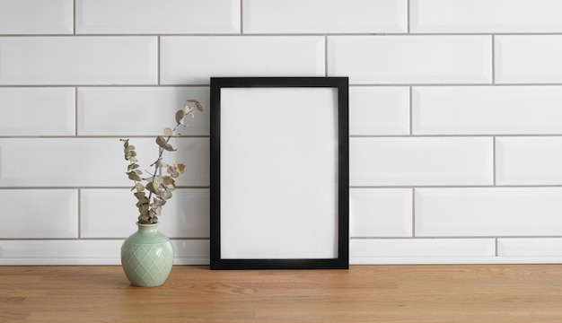 Een frame met een leeg doek tegen een witte tegelmuur en op een houten tafel met vaas