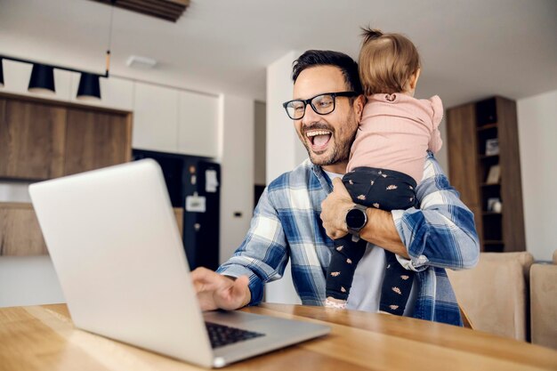 Een freelancer probeert zijn werk op afstand af te maken op een laptop en houdt zijn dochtertje in zijn armen