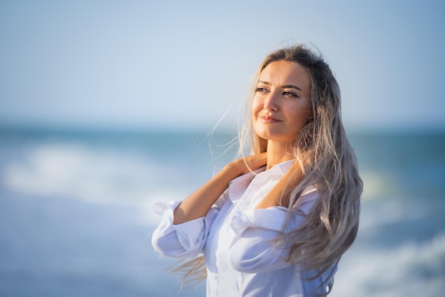 Een gebruind meisje met lang blond haar in een delicaat blauwachtig zwempak en een wit licht overhemd gaat door haar lange haar aan de kust van een stormachtige zee