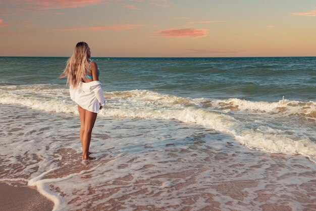 Een gebruind slank meisje in een blauwachtig zwempak en een wit licht overhemd geniet van een warme zonnige zomer aan de kust van een razende blauwe zee