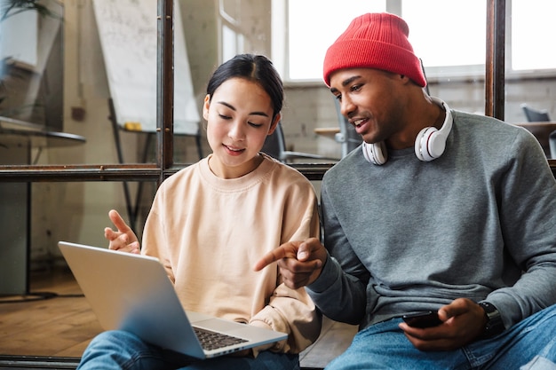 Een geconcentreerde collega's werken binnenshuis op kantoor met een laptopcomputer met behulp van een mobiele telefoon.