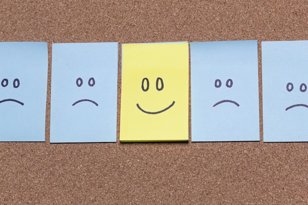 Foto een geel kleverig briefje met een gelukkig gezicht steekt uit een rij blauwe briefjes met verdrietige gezichten
