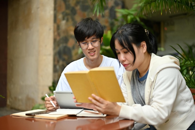 Een gefocuste Aziatische vrouwelijke universiteitsstudent leest een boek terwijl haar mannelijke vriend een tablet gebruikt