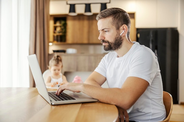 Een gefocuste externe medewerker typt op een toetsenbord op een laptop terwijl hij op zijn dochter past