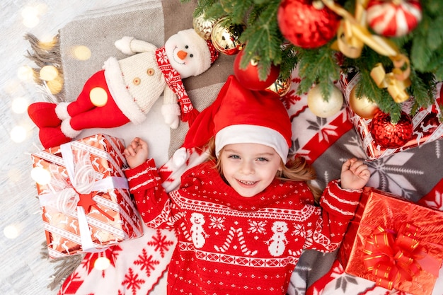 Een gelukkig babymeisje ligt thuis onder de kerstboom en wacht op het nieuwe jaar of Kerstmis in een rode kerstmanhoed en glimlacht en verheugt zich