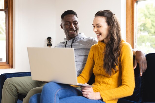 Een gelukkig, divers koppel zit op de bank met een laptop thuis.