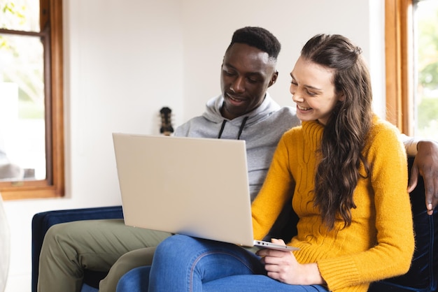Een gelukkig, divers koppel zit op de bank met een laptop thuis.