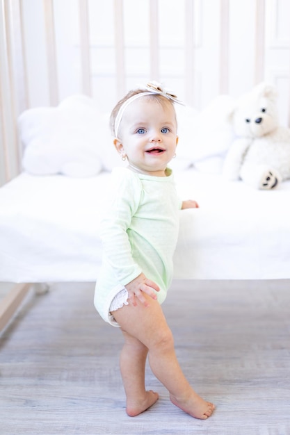 Een gelukkig en glimlachend klein babymeisje staat of klimt op de wieg in de kinderkamer in een wit katoenen rompertje dat de baby leert lopen