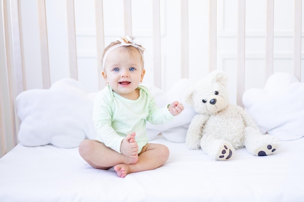 Een gelukkig en glimlachend klein babymeisje zit in een wieg in een kinderkamer in een witte katoenen bodysuit met teddyberen en lachen