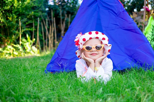 Een gelukkig en vrolijk kind een meisje ligt op het groene gras bij de tipi op de speelplaats in een zonnebril in de zomer en glimlacht