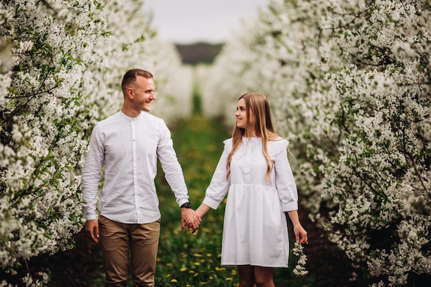 Een gelukkig jong verliefd stel staat in een tuin met bloeiende appelbomen. Een man in een wit overhemd en een meisje in een witte lichte jurk lopen in een bloeiend park