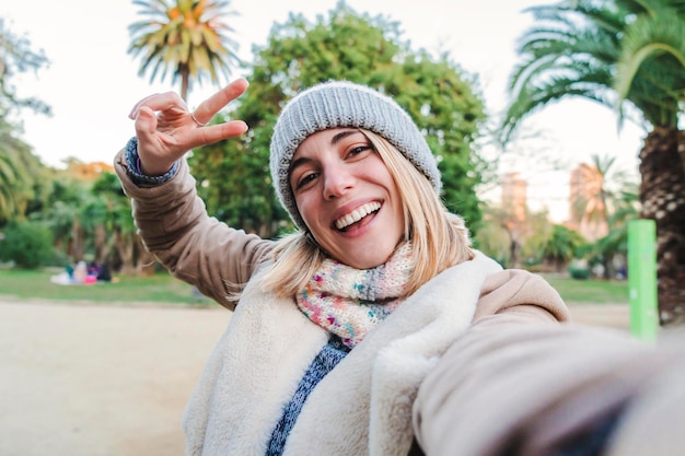 Foto een gelukkig kaukasisch blond meisje glimlachend een selfie-portret nemen met een mobiele telefoon europese gelukkige jonge vrouw die het vredesteken doet met de vingers in een park buiten lifestyle-concept